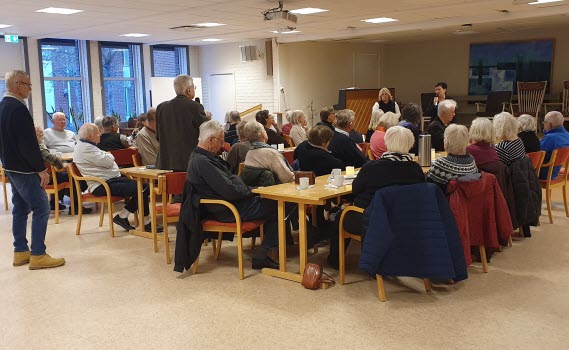 seniorer vid långbord i samlingssal. En person står upp och ställer fråga i en mikrofon, två föredragshållare står redo att svara på frågan