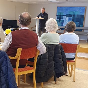 Man vid en talarstol och i förgrunden del av en sittande publik