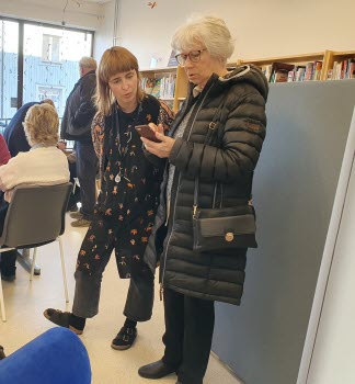 Två damer som står i en bibliotekslokal. En av dem har en mobiltelefon i handen