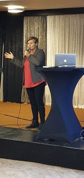Kvinna på scen, pratar i mikrofon och sträcker ut en hand i gest mot publiken