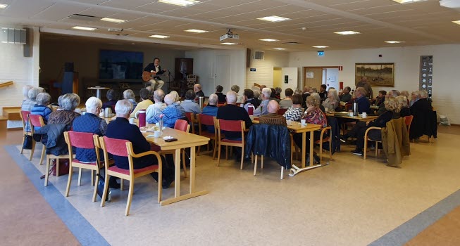 Samlingssal med ett 50-tal pensionärer som lyssnar till en trubadur på en scen