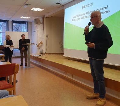En man håller i en mikrofon. Han hälsar två föredragshållare välkomna. De väntar i bakgrunden och mellan dem visas en storbild via projektor.