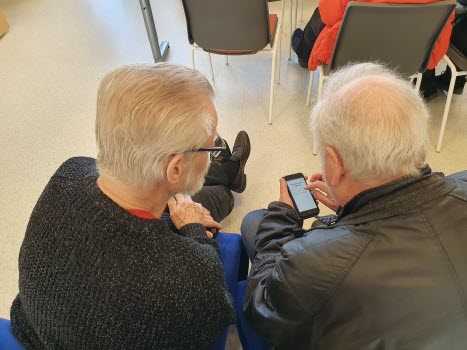 två seniorer fotograferade bakifrån. En av dem har en mobiltelefon i handen