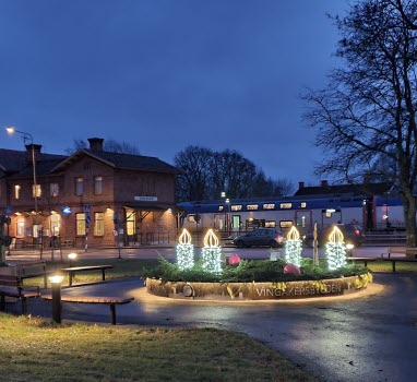 Järnvägsparen i Vingåker. Fyra ljus lyser i en rundel med granris. I bakgrunden stationshuset och ett tåg som gjort ett stopp