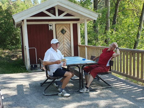 Man och kvinna fikar vid campingbord. Friggebod i bakgrunden.